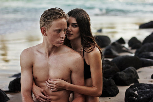 A young couple in a close embrace on the beach, with the man shirtless and the woman wearing a black top, set against a calm ocean and rocks in the background.