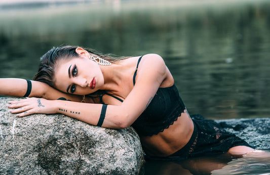 Model posing on a rock by the water, wearing black lace attire, with bold makeup and 'ROSE GANG' tattoo visible on her arm